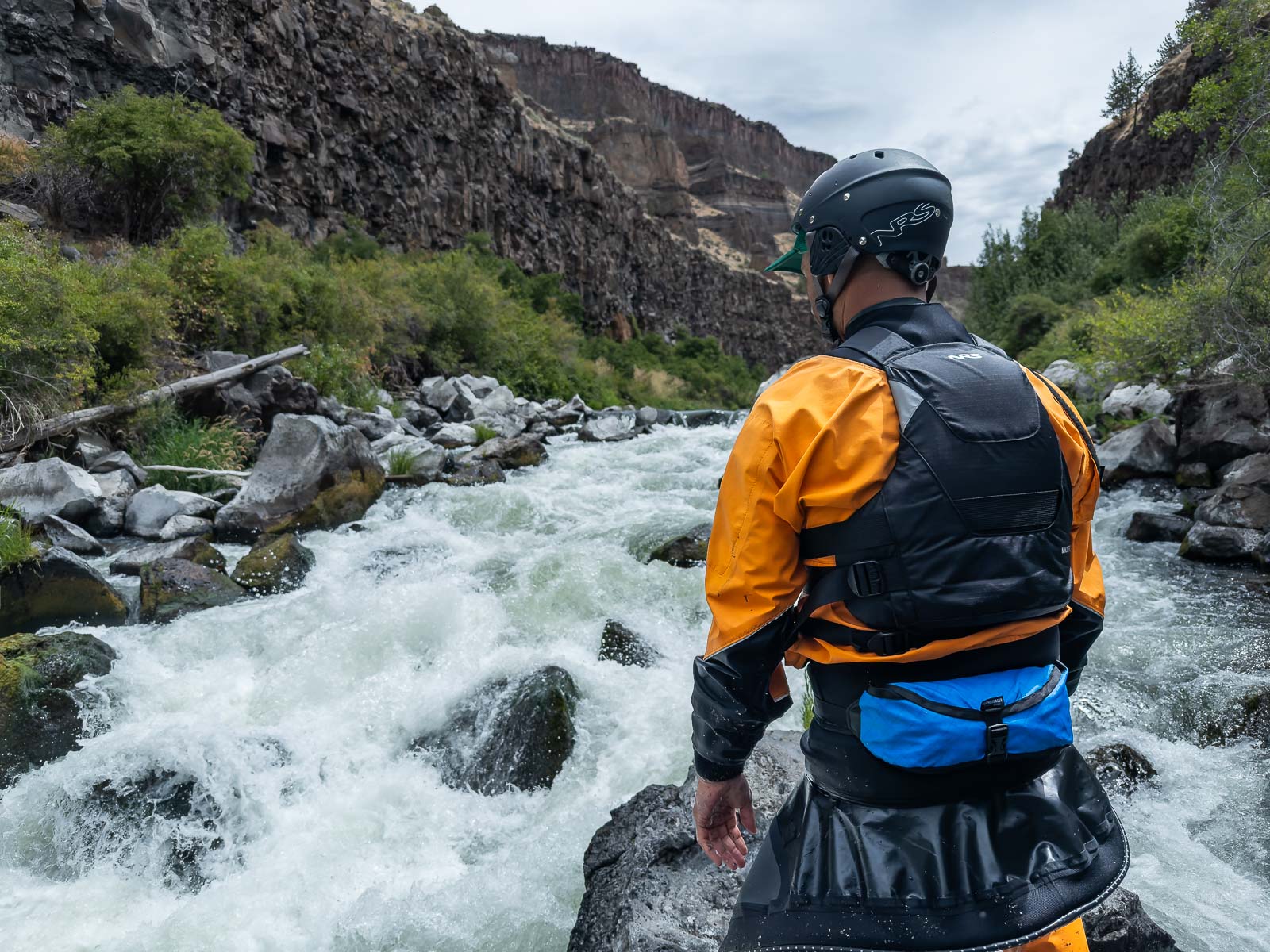 Person in whitewater gear with a Sockdolager Flare Throw Bag standing by a river with rapids and mountains in the background.