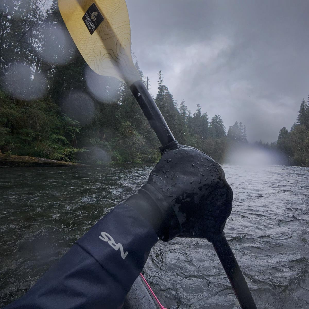 Wearing pogies and paddling a Packraft on a misty river with trees lining the banks